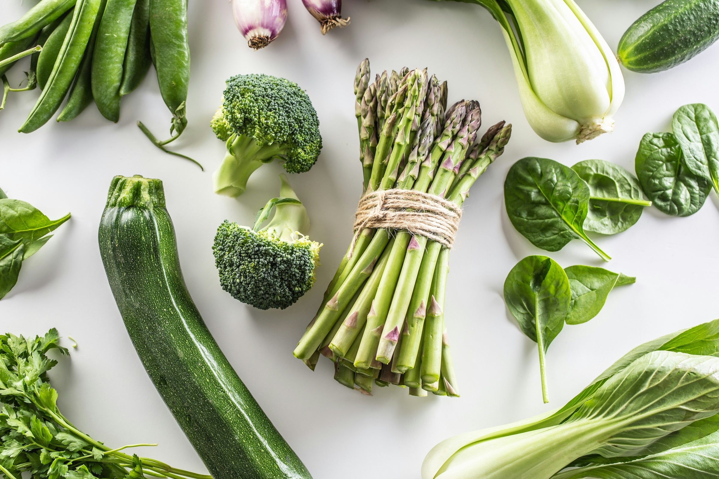 Frisches grünes Gemüse, darunter Brokkoli, Zucchini, Spargel, Spinat, Pak Choi und Erbsen, auf einem weißen Hintergrund arrangiert.